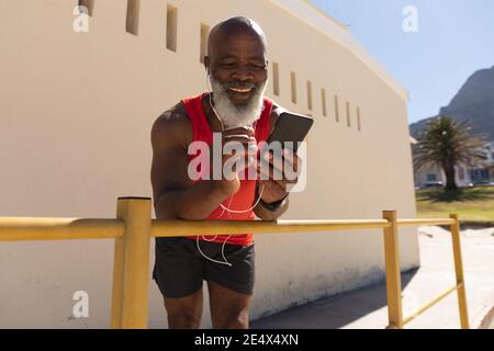 Fit senior african american man wearing earphones using smartphone in sunny street smiling. healthy retirement technology communication outdoor fitnes Stock Photo