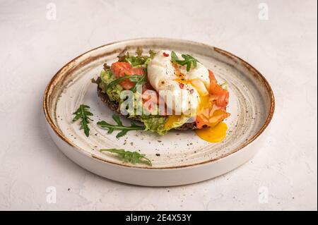 Homemade poached egg toast with avocado, salted salmon, arugula and rye bread on a ceramic plate, close up. Healthy food concept Stock Photo