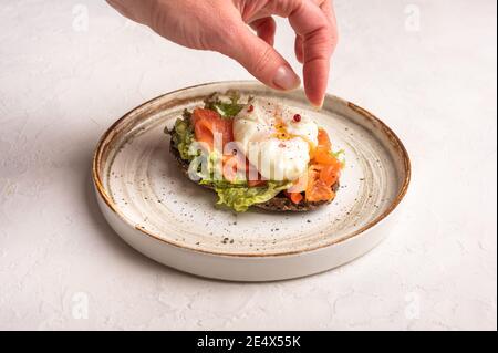 Woman's hand puts pepper on poached egg toast with avocado, salted salmon, arugula and rye bread on a ceramic plate, close up. Healthy food concept Stock Photo