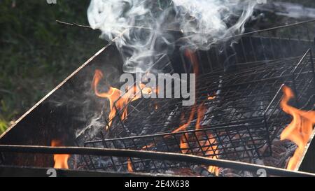 Fire burning in grill with old metal grid with ashes and grey flying smoke. Flames and smoke from rustic mangal outdoors Stock Photo