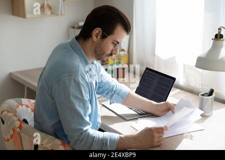 Concentrated young man remote employee doing paperwork by laptop screen Stock Photo