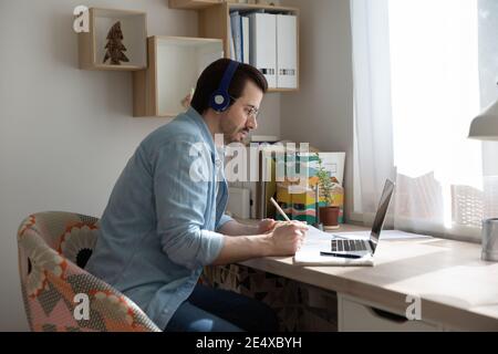 Attentive male student listening to lecture online using earphones laptop Stock Photo