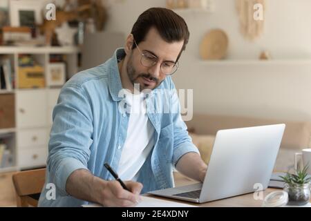 Concentrated man sitting by computer taking notes of distant lecture Stock Photo