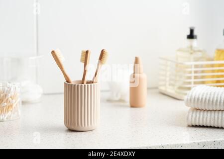 Cup with bamboo toothbrushes and stack of cotton towels placed on shelf near hygienic supplies in morning in bathroom Stock Photo