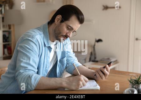 Young male student watching distant lecture by smartphone making notes Stock Photo