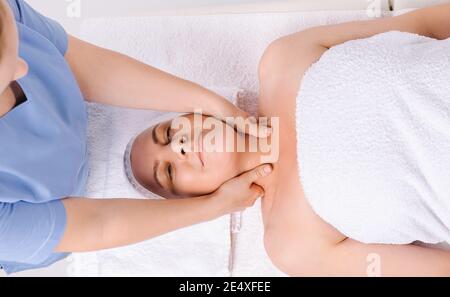 The master masseur is giving a relaxing head and neck massage to an older woman wrapped in a white towel. Stock Photo