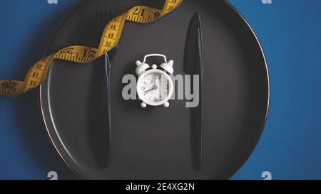 Flat lay fork and kitchen knife on a plate with tape measurement and white alarm clock. Intermittent fasting concept dieting healthy lifestyle