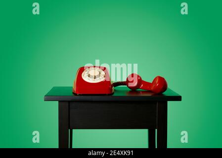 a red landline rotary dial telephone, with its handset off the hook on a black wooden table, on a green background Stock Photo