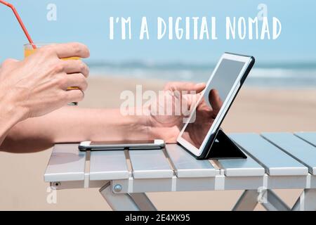 closeup of a young caucasian man on the beach, sitting at an outdoors table, using a digital tablet, while is having a refreshing orange beverage, and Stock Photo