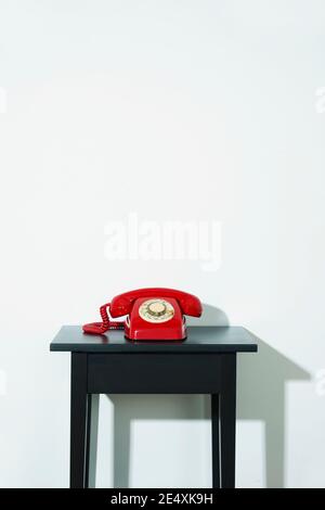 a red landline rotary dial telephone on a black wooden table, indoors, in front of a white wall with some blank space on top Stock Photo