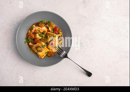 Traditional Sicilian fish with tomatoes, olives, parsley and red pepper in a deep plate with fork on a light background, close up Stock Photo