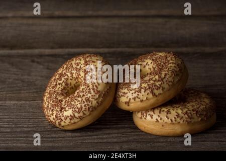 Yummy cake donuts are lying on a wooden background. Three donuts are covered with vanilla glaze. They are all topped with sprinkles. Stock Photo