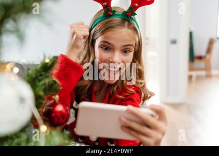 Excited caucasian girl making winner gesture while playing video game on cellphone at home Stock Photo