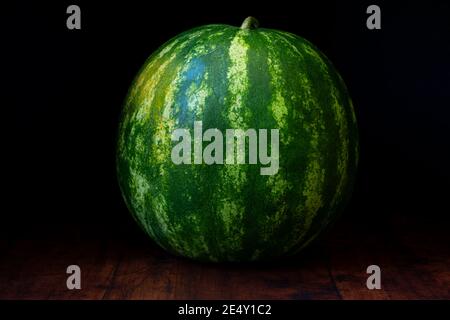 Watermelon fruit is lying on a rustic wooden background. This juicy beauty is emerging from the dark and concealing refreshing sweetness. Stock Photo