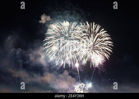 Bright white fireworks lights in the dark sky. Small city celebrates new year with colorful fireworks at night. Gray firework smoke cloud in the sky. Stock Photo