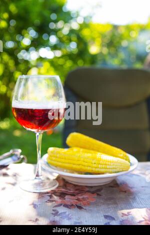 A glass of dark beer in a glass along with a plate of boiled corn, standing on the table. Summer picnic, beautiful sunshine Stock Photo