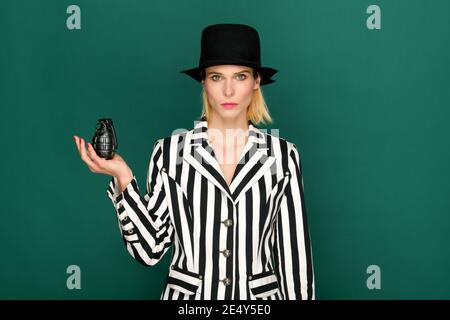 Confident stylish blond model in black and white striped jacket and hat holding black grenade on green background Stock Photo