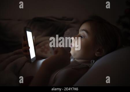 A cute little girl lies on a pillow and enthusiastically plays on the phone at night in bed instead of sleeping. Stock Photo