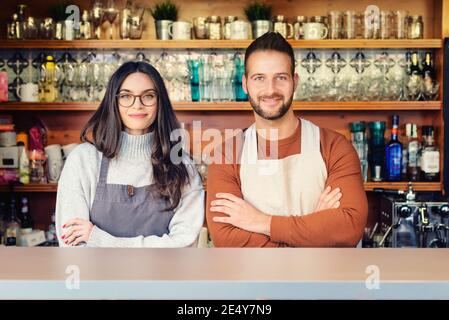 Shot of young waitress and waiter standing behind the counter in the cafe while waiting for their guest. Small business. Stock Photo