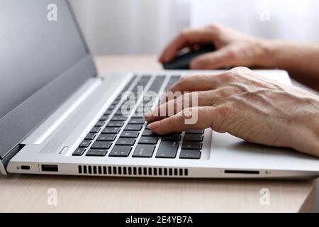Elderly woman working at the laptop. Wrinkled female hands on notebook keyboard on a desk in sunlight Stock Photo