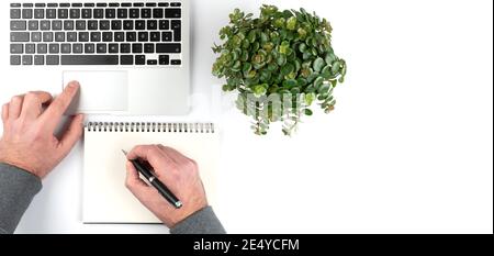 overhead view of person taking notes on notepad white working on laptop computer on white desk with green potted plant Stock Photo