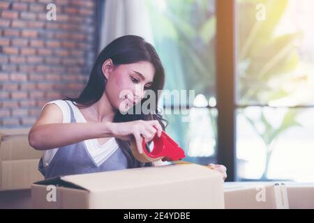Businesswoman taping up a cardboard box, Asian woman packing boxes among stack of parcels in her shopping online business at home Stock Photo