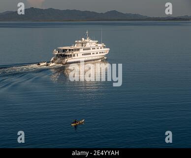 The Australian mega yacht True North cruises past the forested hills of Bougainville, Papua New Guinea, a man paddling alone in a narrow canoe in the foreground. Stock Photo