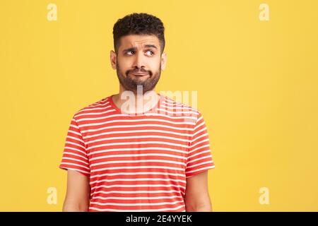 Doubtful bearded man in striped t-shirt standing with thoughtful expression, remembering or planning, thinking over answers. Indoor studio shot isolat Stock Photo