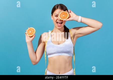 Smiling active woman in white sportswear with tape line on shoulders closing eye with grapefruit half, having fun and losing extra weight on fruits di Stock Photo