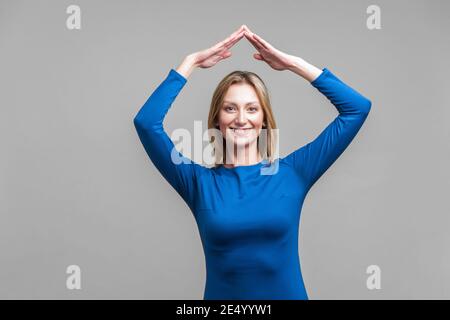 Secured life, insurance concept. Portrait of cheerful woman in elegant tight blue dress showing home roof gesture with hands over head, feels safe. in Stock Photo