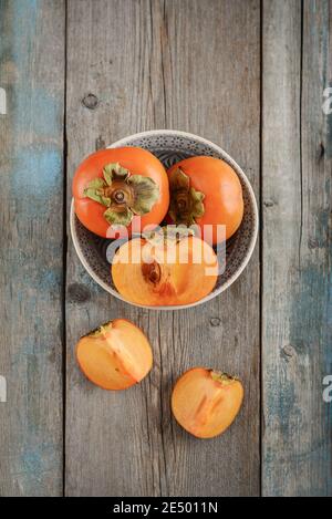 Fresh organic ripe Persimmon fruits in bowl with slice on old wooden background, top view Stock Photo