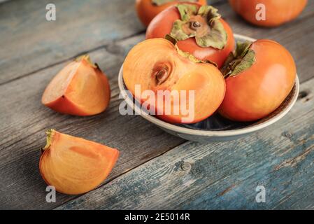 Fresh organic ripe Persimmon fruits in bowl with slice on old wooden background. Stock Photo