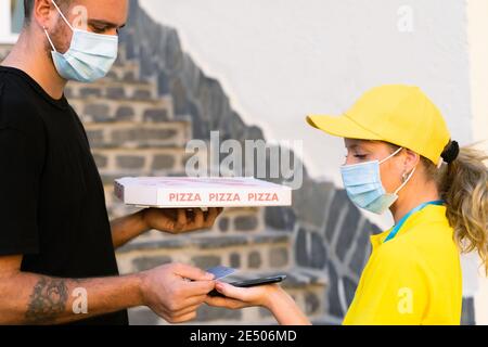 Delivery woman employee in yellow cap and t-shirt uniform face protective mask. Man hold a pizza box and pay with credit card. Coronavirus and buy con Stock Photo