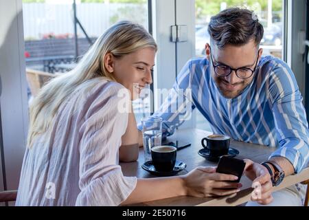 Young romantic couple enjoying a cup of coffee together in a cafe, sitting at the table and talking while scrolling through social media on the phone. Stock Photo
