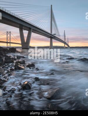 Bottom view of a large three-tower cable-stayed bridge at sunrise. In the foreground, sea waves rolling on the stones of the coast. Queensferry Crossing Bridge, Scotland, United Kingdom. Stock Photo