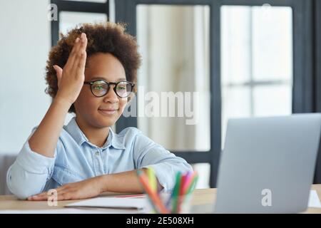 Be engaged. Enthusiastic mixed race teen schoolgirl in glasses raising her hand during online video lesson while studying from home Stock Photo