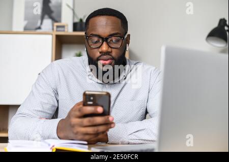 Serious young African-American adult in glasses sitting at the desk, listening to the music on his mobile phone, working on social media projects, and Stock Photo