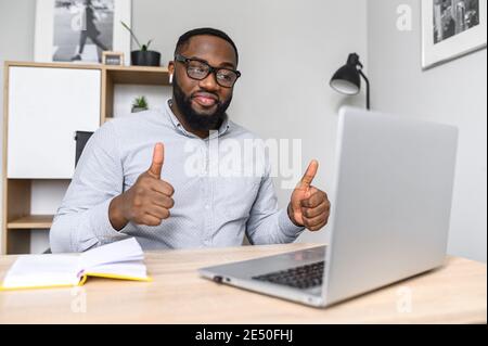 An intelligent young African-American teacher approves the project online on the laptop, wearing glasses, making notes, showing thumbs-up. An excited Stock Photo