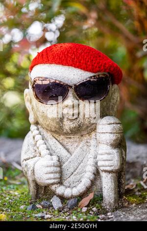 Close up of a small stone japanese jizo statue wearing a red wool cap and sunglasses against a green bokeh background Stock Photo