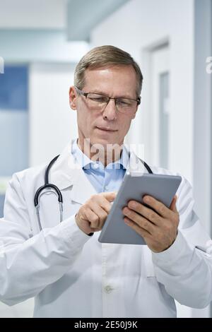 Mature doctor wearing glasses using digital tablet in hospital. Stock Photo