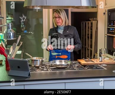 Angela Berg stands in a black chef's jacket bearing her name at the stove of her kitchen in Grevenbroich, Germany, an iPad showing the live video stream in front of her on the kitchen island. She holds the packaging of the smoked salmon trout "La Regina di San Daniele", an empty pan and a pot stand ready. The Michelin-starred restaurant Harry's Piccolo in Trieste, Italy, remains closed to guests during the coronavirus pandemic and sells cooking boxes with a live video class to private households instead. Angela Berg takes part in one of these classes this evening, led by the restaurant's chef  Stock Photo