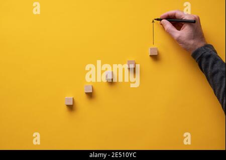 Male hand drawing an upward pointing arrow on top of a growing graph made of wooden blocks. Over yellow background. Stock Photo