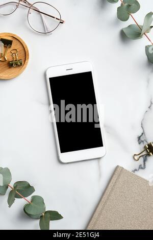 Home office workspace with smartphone mockup, feminine glasses, notebook, eucalyptus leaves. Flat lay, top view, copy space. Stock Photo