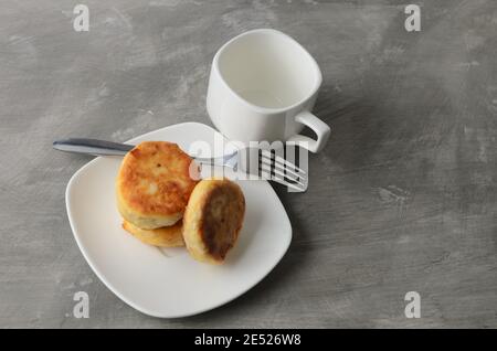 A few pancakes on a white plate with a fork and an empty mug on a textured gray background. Selective focus. Stock Photo