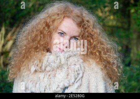 Portrait of a beautiful reddish blond woman in her forties with wild red curls stands in the green forest, copy space Stock Photo
