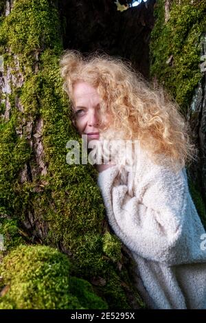 redheaded woman in her fifties with a fur collar snuggles up against a tree trunk overgrown with moss. Stock Photo