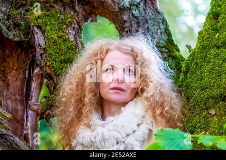 redheaded woman in her fifties with a fur scarf collar stands in the forest overgrown with moss. Stock Photo