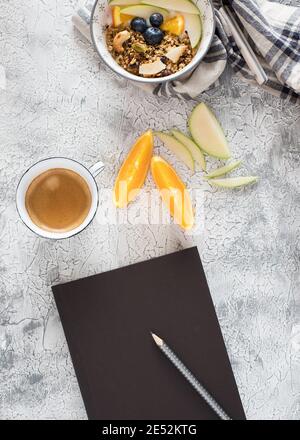 Empty opened black notebook with a pencil over it, a granola bowl at the top and a foamy coffee cup with some orange and apple slices in the middle. Stock Photo