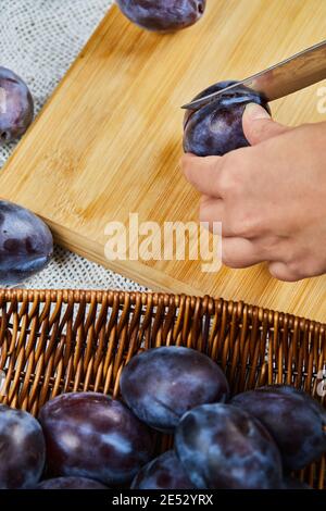 Cutting fresh plum on wooden board with basket of plums Stock Photo