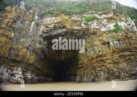 Catlins New Zealand - February 24 2015; Tourist walking into  weathered rock face  entrance to black hole of cave Cathedral Caves on Waipati Beach ris Stock Photo